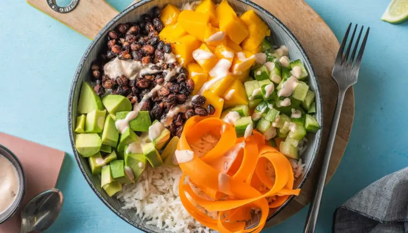 En färgglad poke bowl med ris, svarta bönor, mango, gurka, avokado och morotsstrimlor, toppad med en krämig dressing.