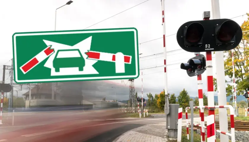 Zielony znak informuje o oznaczeniu przejazdu kolejowego. Po bokach widać rogatki i sygnalizację świetlną.