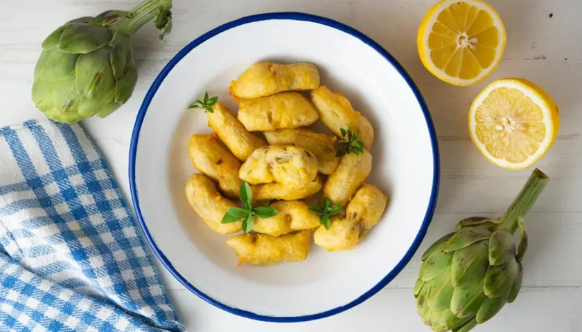 Carciofi fritti dorati, preparati con una pastella croccante, serviti in un piatto bianco con limone e carciofi freschi.