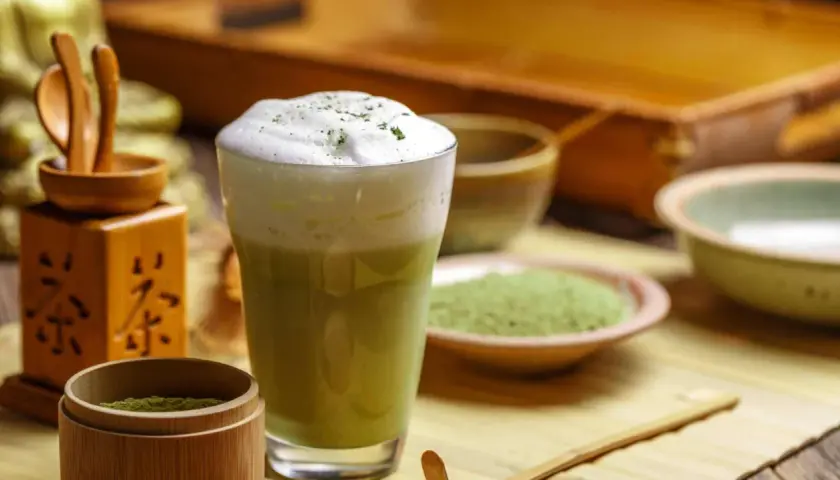 Ein cremiger Matcha Latte mit Milchschaum, umgeben von Utensilien für die Matcha Tee Zubereitung.