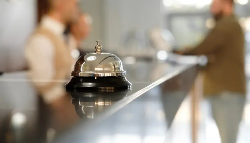 Dzwonek hotelowy na ladzie, symbol usług uzupełniających. W tle niewyraźne postacie personelu.