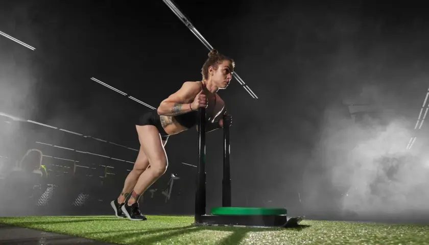 Kobieta z tatuażami pcha sanie treningowe. To właśnie crossfit co to – siła, wytrzymałość i determinacja w akcji.