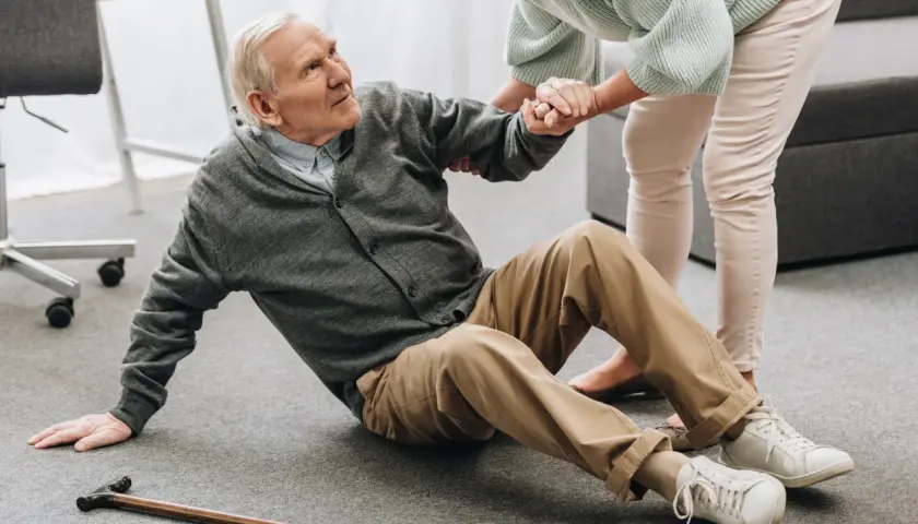 Älterer Mann sitzt auf dem Boden, wird von einer Frau gestützt. Ein Stock liegt daneben. Dies illustriert die Notwendigkeit von Sturzprophylaxe.