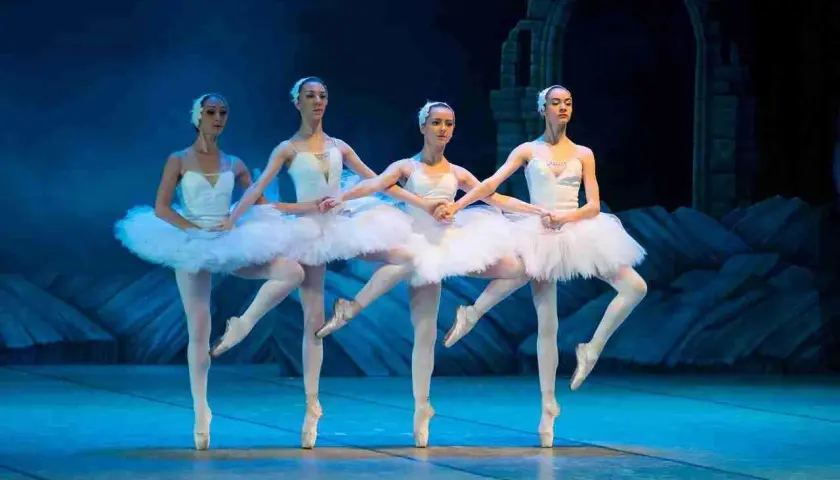 Quattro ballerine in tutù bianchi eseguono una danza aggraziata sul palco, con un arco di pietra sullo sfondo.