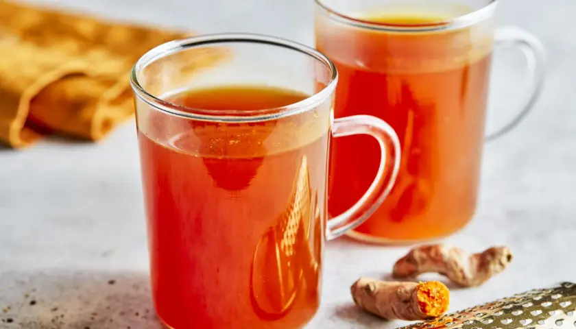 Zwei Gläser mit leuchtend orangefarbenem Getränk, das nach einem Kurkuma Honig Rezept zubereitet wurde. Frische Kurkumawurzeln und ein Sieb liegen daneben.