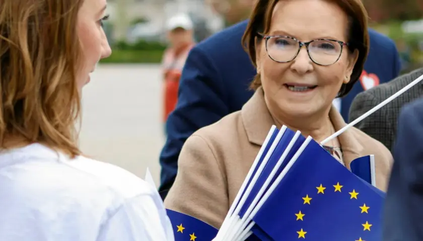 Ewa Kopacz, gdzie mieszka, uśmiecha się, trzymając flagi UE.