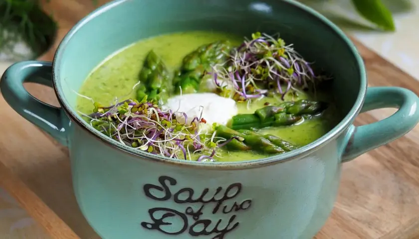 Aksamitna zupa z zielonych szparagów, udekorowana kiełkami i kleksem śmietany, w ceramicznej misce z napisem "Soup of the Day".