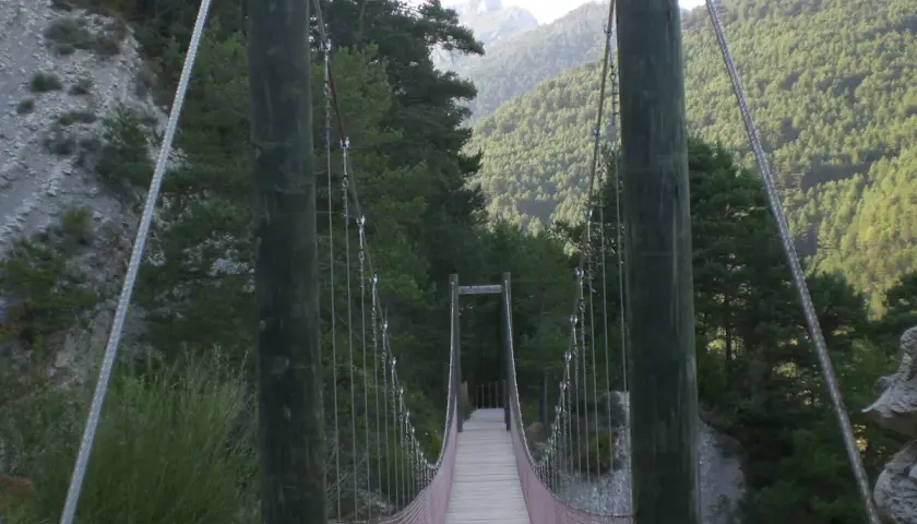Puente colgante de madera y metal en la vía del Nicolau, rodeado de bosques y montañas.