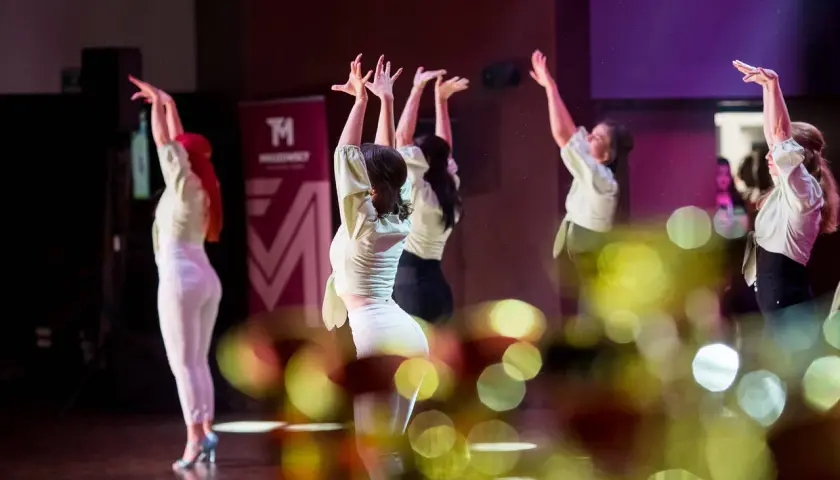 Tancerki w jasnych bluzkach i białych spodniach wykonują zmysłowy taniec. W tle logo szkoły tańca. To piękna **bachata solo**.