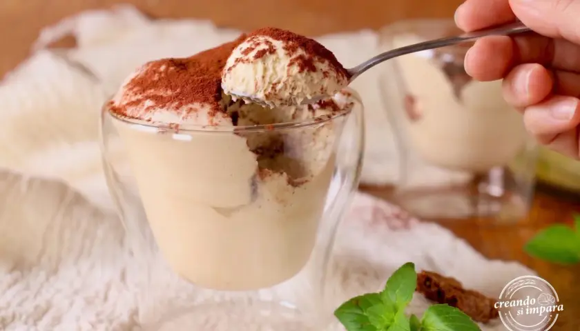 Un cucchiaino preleva una porzione generosa di soffice mousse al caffè, cosparsa di cacao, da un bicchiere di vetro.