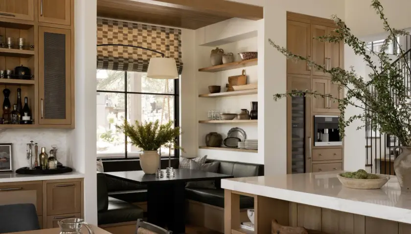 A cozy breakfast nook built in with a dark table, plush seating, and a large window. Shelves display rustic pottery and kitchenware.