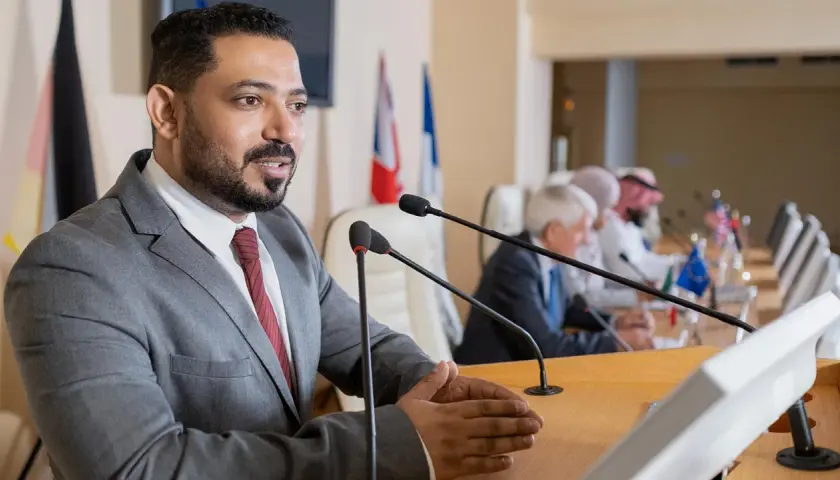 A man speaks at a podium, perhaps sharing insights from his pursuit of best online MPA programs, as others listen at a long table with flags.