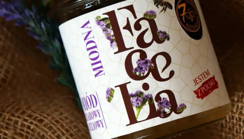 Słoik z miodem faceliowym, ozdobiony fioletowymi kwiatami i pszczołą. Etykieta głosi "Miodna Facelia. Jestem z Polski".