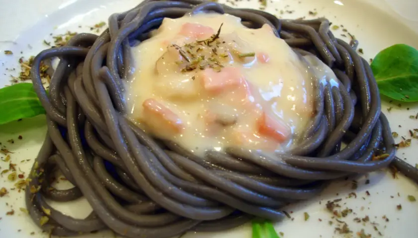 Espaguetis negro con nata, trocitos de jamón y hierbas aromáticas, servido en un plato blanco.