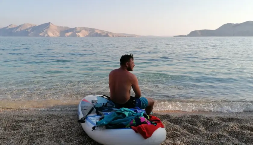 Chorwacja: mężczyzna na desce SUP na kamienistej plaży, w tle góry i spokojne morze. Idealne miejsce na relaks.