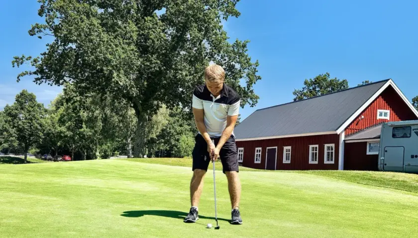 Man puttar på en golfbana, en röd byggnad och träd i bakgrunden. Golf som friskvård är en härlig aktivitet.