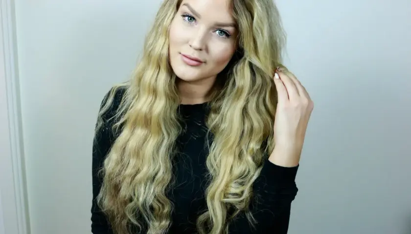 En ung kvinna med långt, blont, vågigt hår som rör vid det. Hon har gröna ögon och ljus hy. Hon bär en svart tröja. Frågan "hur får man lent hår" kan besvaras med denna look.