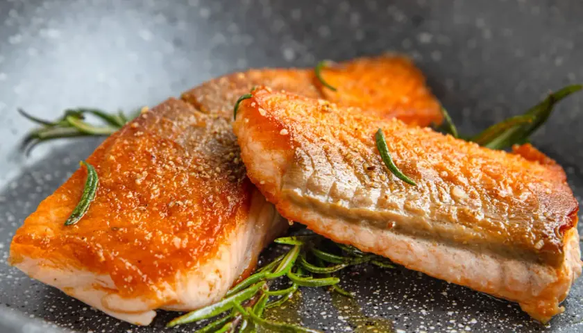 Zwei perfekt gebratene Lachsfilets mit knuspriger Haut und Rosmarin in einer Pfanne.