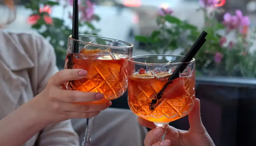 Dwie osoby wznoszą toast Aperol Spritz, ozdobionymi owocami i słomkami, na tle kwitnących kwiatów.