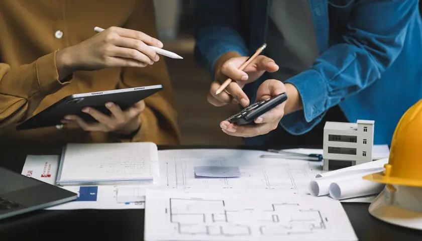 Architekci analizują plany, liczą koszty i omawiają, ile kosztuje odbiór domu. Na stole szkice, tablet, kalkulator i model budynku.