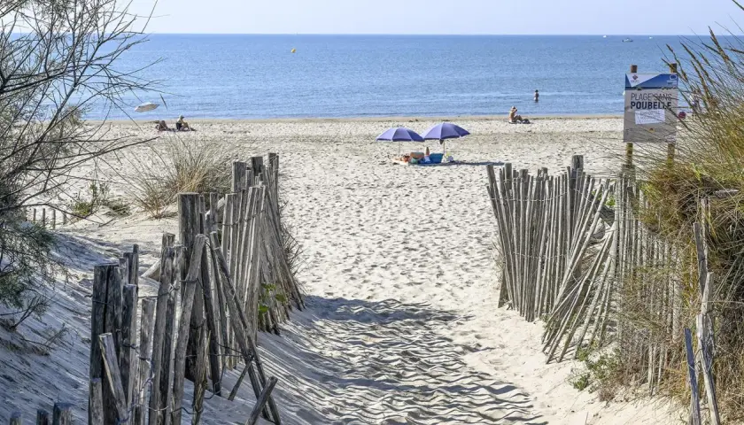 Ein sandiger Weg führt zum Montpellier Strand. Zwei Sonnenschirme spenden Schatten, während Menschen die Sonne genießen.