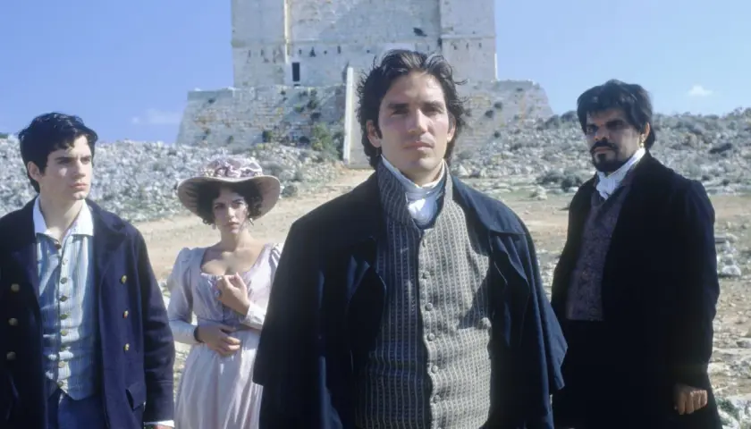Młody mężczyzna w stroju z epoki, jak z filmu hrabia Monte Cristo 2024, stoi na tle kamiennej budowli. Obok niego kobieta w kapeluszu i inny mężczyzna.