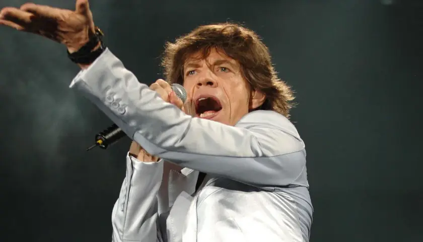 Mick Jagger śpiewa, wykonując swoje legendarne utwory. Wokalista w srebrnej marynarce z mikrofonem w dłoni.