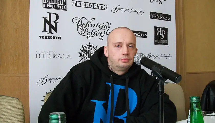 Peja w czarnej bluzie z niebieskim logo, przed mikrofonem. Na ścianie za nim napisy: "TERRORYM", "REEDUKACJA", "Definicja Peja". Wiecie co z nim zrobić.
