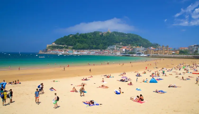 Gente disfrutando de la arena dorada y el mar turquesa en la que podría ser la mejor playa de España, con la ciudad y un monte verde de fondo.