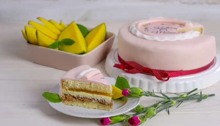 Różowy tort z napisem "Kocham Mamę", udekorowany bielą tytanową spożywczą, obok kawałek ciasta i mango.