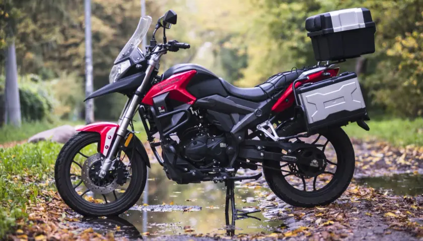Czerwono-czarny motocykl szosowo turystyczny 125 z kuframi, gotowy na jesienne wyprawy po leśnej drodze.