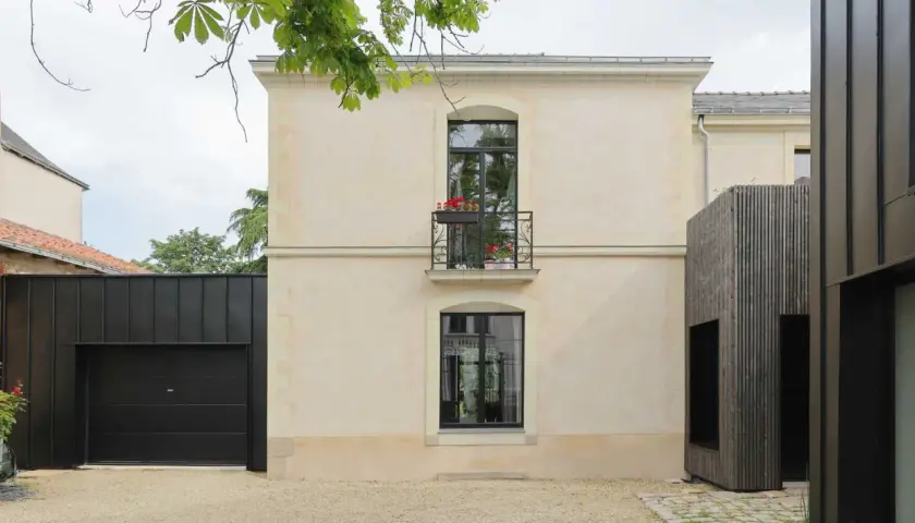 Maison rénovée par un architecte DPLG, mêlant pierre claire, bois sombre et garage noir moderne. Balcon fleuri sur la façade.