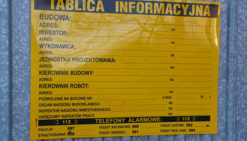 Tablica informacyjna na budowie. Widać pola na dane inwestora, kierownika budowy, numer pozwolenia na budowę i telefony alarmowe. Ważne jest pozwolenie na budowę, by wszystko było legalne.