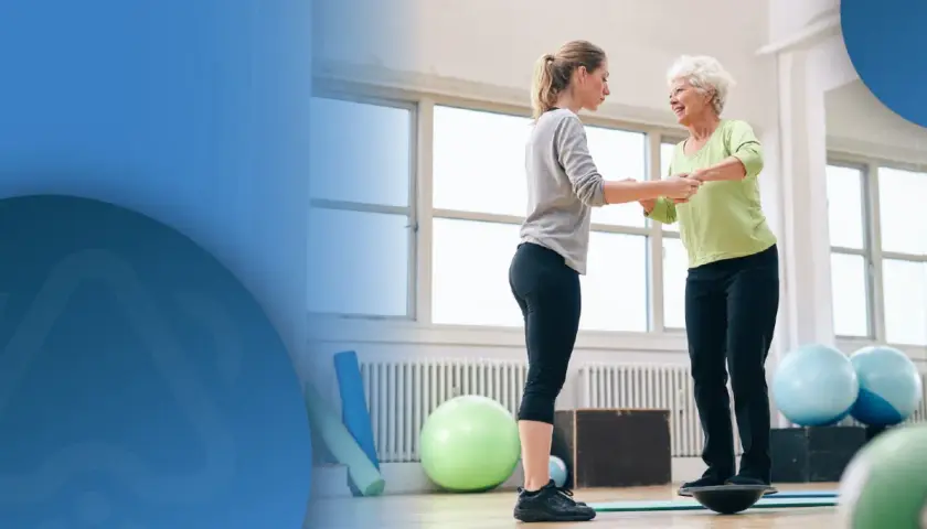 Physiotherapeutin hilft älterer Frau auf Balance Board, um Sturzgefahr zu reduzieren.