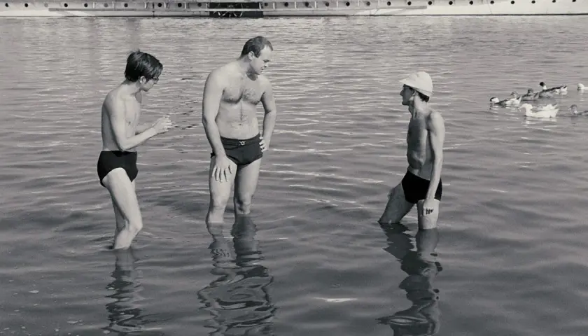 Trzech mężczyzn w kąpielówkach stoi w wodzie. Jeden z nich, Stanisław Tym w filmie Rejs, rozmawia z kolegami.
