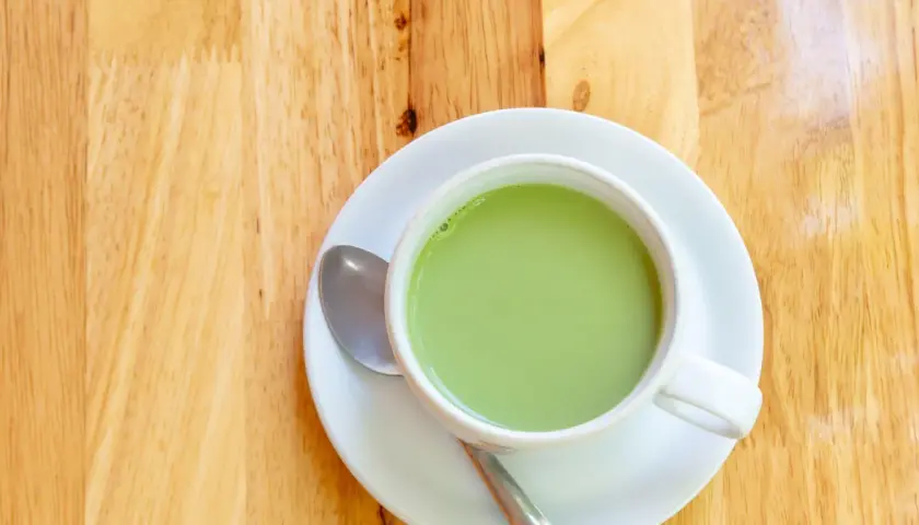 A creamy, vibrant zielona herbata z mlekiem w białej filiżance na drewnianym blacie. Obok leży łyżeczka.