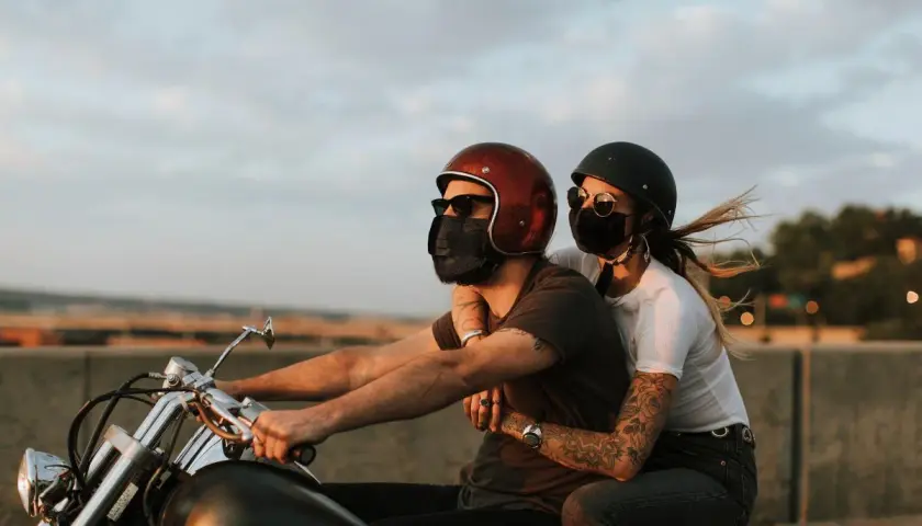 Pareja con mascarillas, montando a horcajadas moto al atardecer. El viento agita el cabello de ella.
