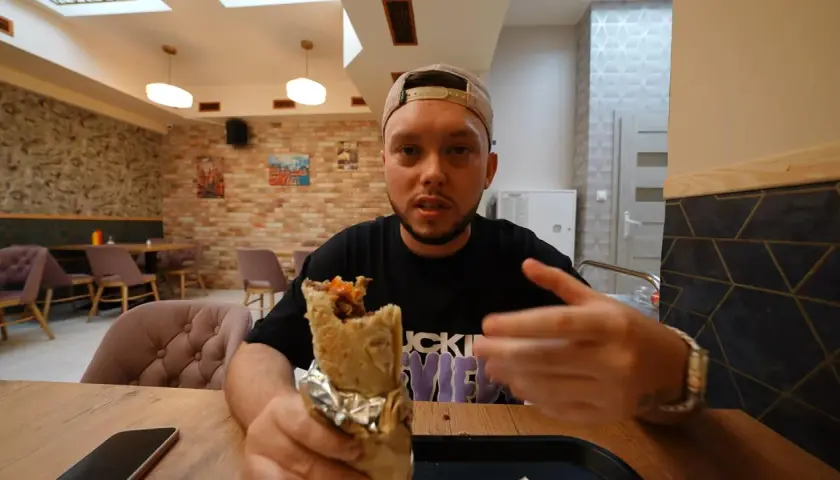 Mężczyzna w czapce z daszkiem trzyma w ręku dużego kebaba, wyglądając jak prawdziwy lider kebab. W tle widać wnętrze restauracji.