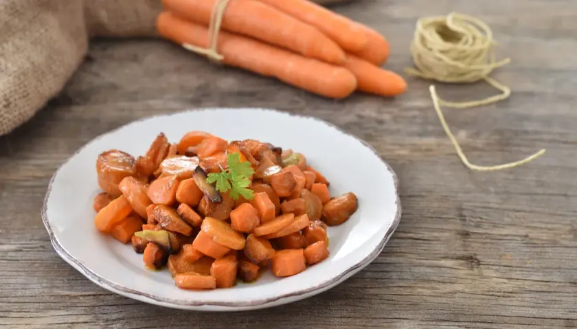 Carote in padella, dolci e caramellate, guarnite con prezzemolo fresco su un piatto bianco. Sullo sfondo, carote fresche legate con spago.