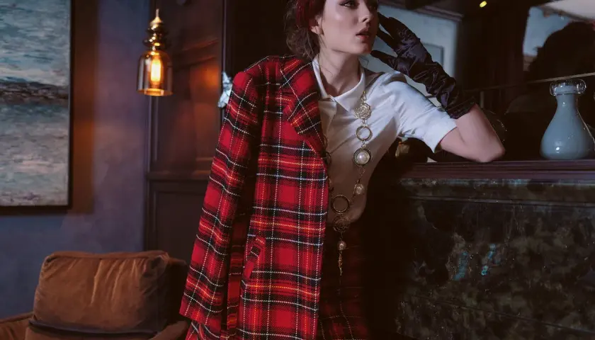 Une femme pose, vêtue d'un blazer au **motif tartan** rouge et noir, avec un collier perlé.