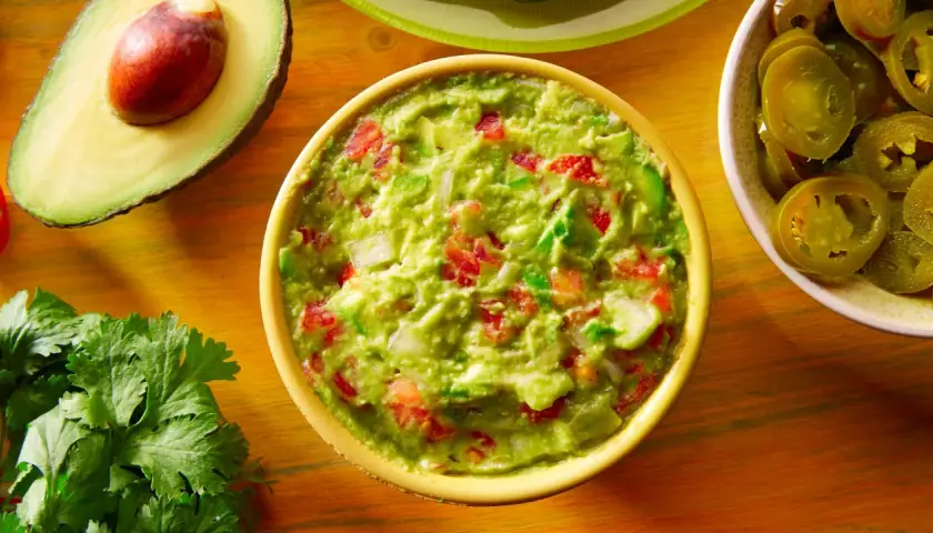 Miseczka guacamole z pomidorami i cebulą, obok awokado, kolendra i plasterki jalapeno.