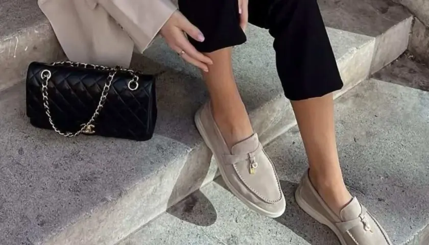 Kobieta w beżowych mokasynach i czarnych spodniach siedzi na schodach obok czarnej torebki Chanel.