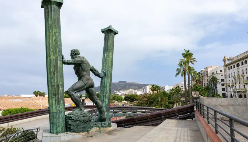 Statua di Ercole che sorregge le colonne d'Ercole oggi, con palme e edifici sullo sfondo.