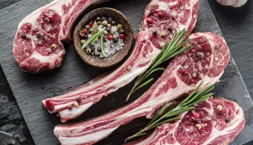 Côtelettes d'agneau crues, assaisonnées de sel et de poivre, prêtes pour une recette d'agneau pascal. Romarin frais et ail complètent la scène.