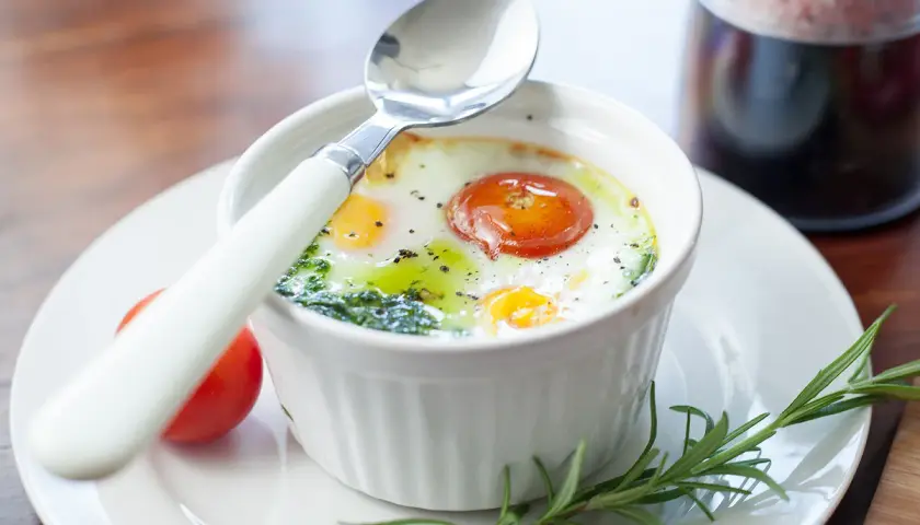 Un œuf ramequin cuit au four, garni d'une tomate et d'herbes, prêt à être dégusté avec une cuillère.