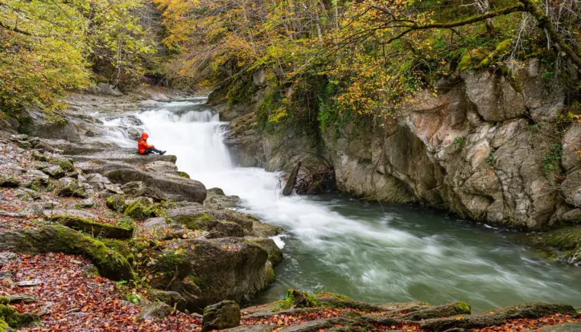 Persona sentada en rocas junto al Saut deth Pish, rodeada de árboles otoñales y agua turbulenta.
