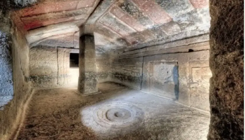 Interno di una tomba etrusca con soffitto dipinto e un pilastro centrale. Un cerchio inciso sul pavimento sembra un antico incantesimo putifigari.