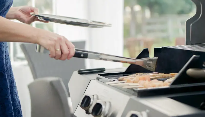 Person i blå klänning använder tång för att vända kycklingspett på en fyrkantig kolgrill.
