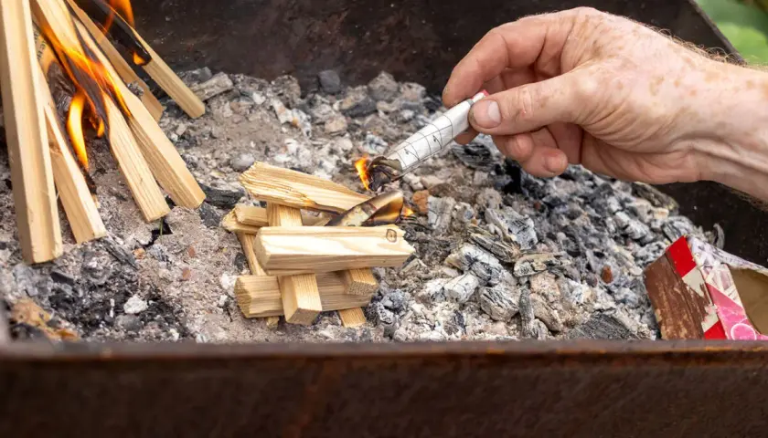 Maintenant, comment allumer un barbecue au charbon : une main allume un morceau de papier roulé pour enflammer des brindilles dans le brasero.