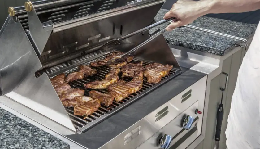 Un homme utilise une pince pour retourner des côtes levées sur un gril à gaz. L'odeur alléchante de la viande cuite emplit l'air.